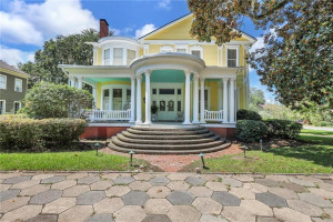 Two-story Victorian-style home at 827 Union Street in Brunswick, GA — pale yellow siding, white-columned front portico with a light blue ceiling, a rounded bay window, red brick foundation and walkway, framed by manicured lawn and mature trees on a bright sunny day