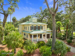 Three-story coastal-style home at 32 Preserve Trail in Waverly, GA — pale yellow siding, green metal roof, white-columned porch and second-floor balcony, a central cupola, and a two-car garage, framed by mature oaks and palm trees on a landscaped lot